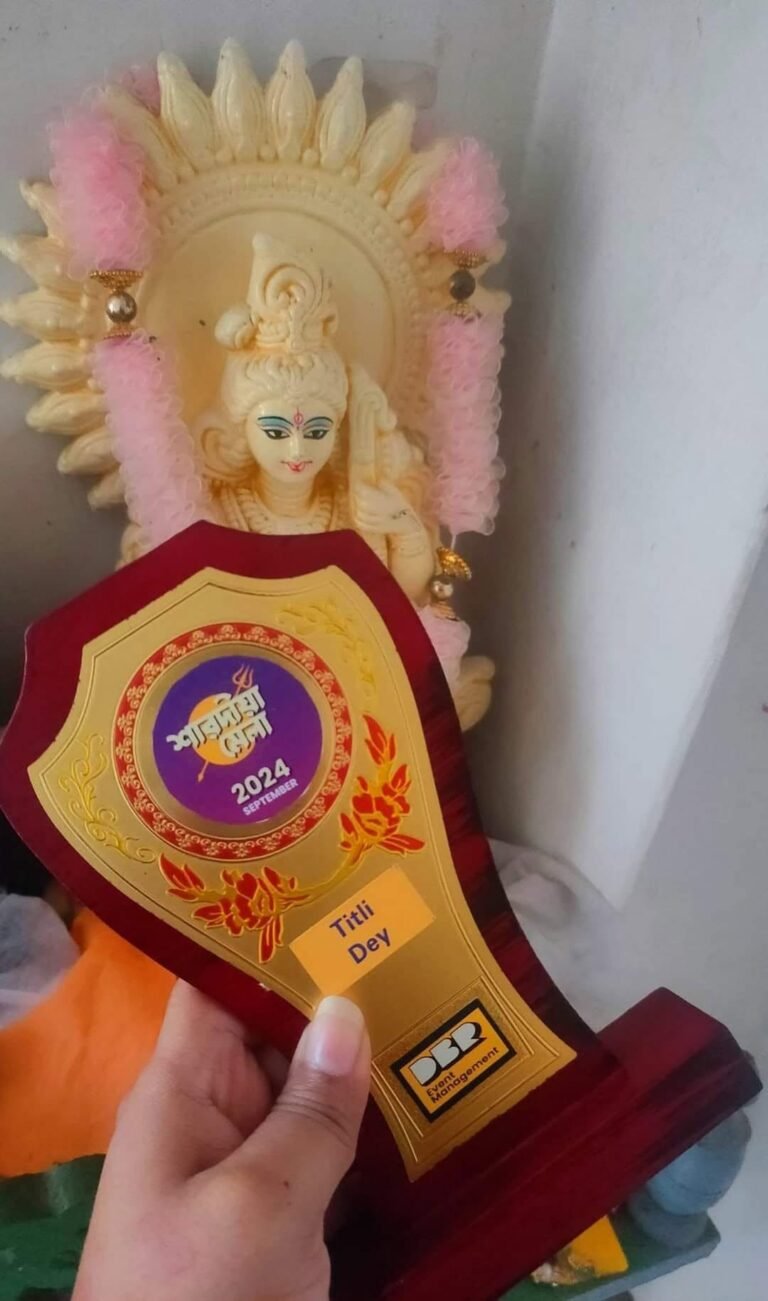 A hand holds a maroon and gold trophy with 'Titli Dey' in front of a white deity idol adorned with pink garlands.