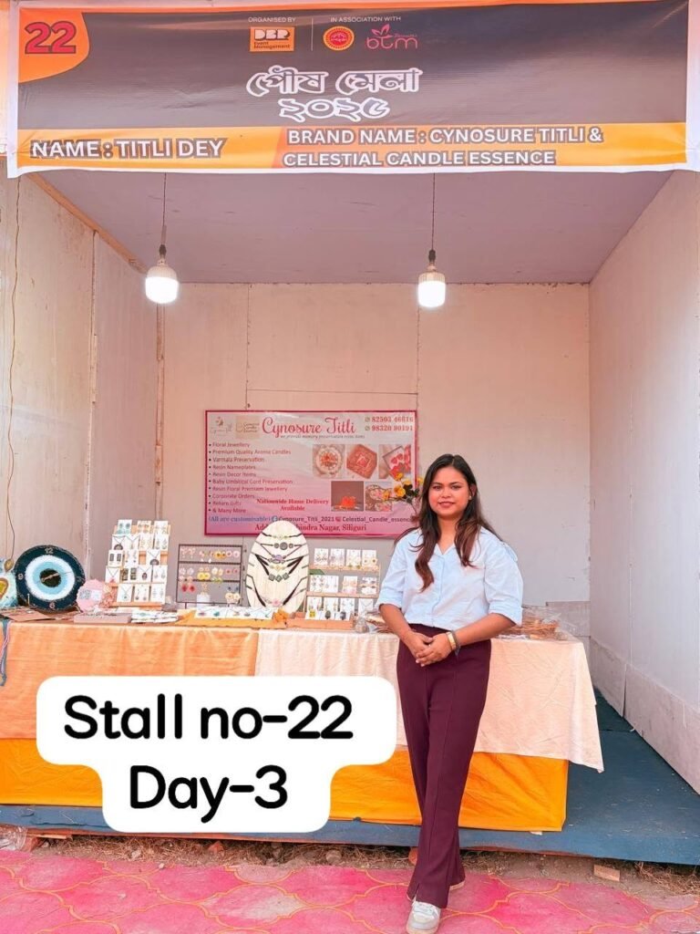 Woman in white shirt and maroon pants standing in front of a stall with handmade resin jewelry, decor, and a banner for Cynos