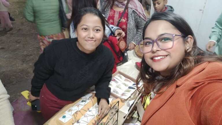 Two women smiling at the camera, standing behind a table displaying handmade resin jewelry and keepsakes at a market stall.