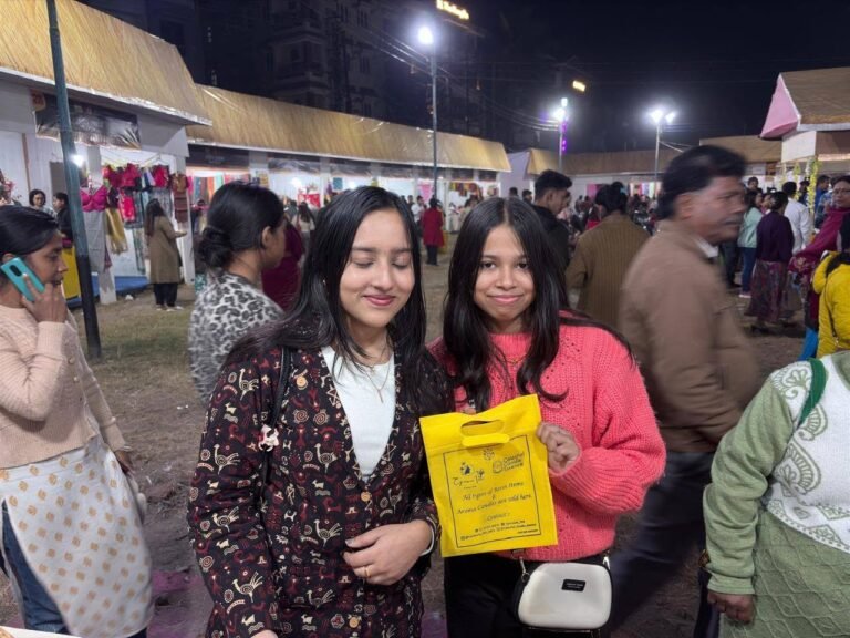 Two smiling women at a night market, one holding a yellow Cynosure Titli bag advertising resin items and aroma candles.