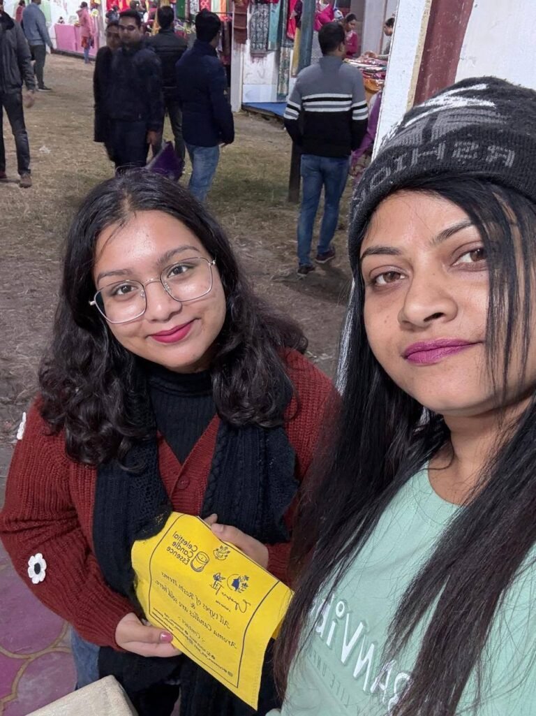 Two women smiling at a fair, one holding a yellow cloth, surrounded by people and stalls.