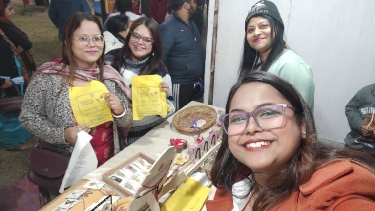 Four smiling women at a market stall with resin jewelry and decor displayed, two holding yellow bags.