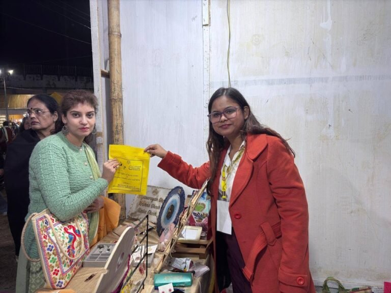 Two women at a market stall, one in a green sweater holding a yellow promotional bag, the other in a red coat.