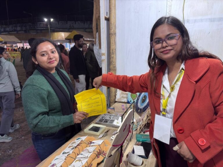 Two women at a market stall with handmade resin jewelry and gifts, one holding a yellow sign, the other smiling in a red coat