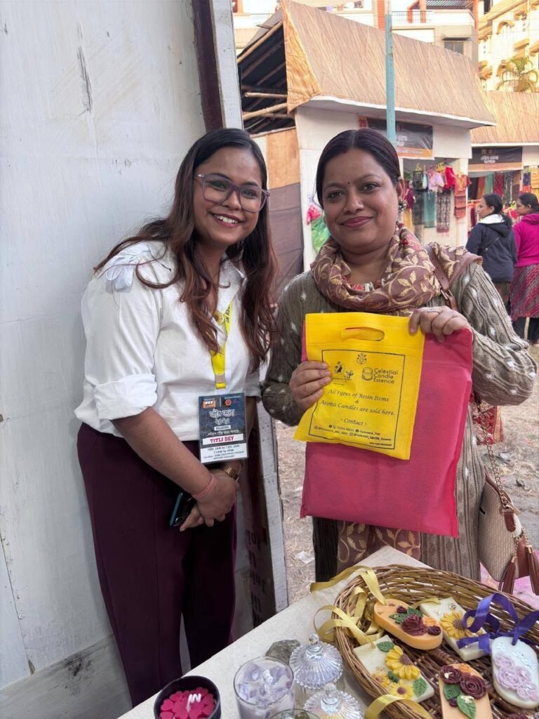 Two women smiling at a market stall with handmade resin items and candles on display, one holding a yellow bag.