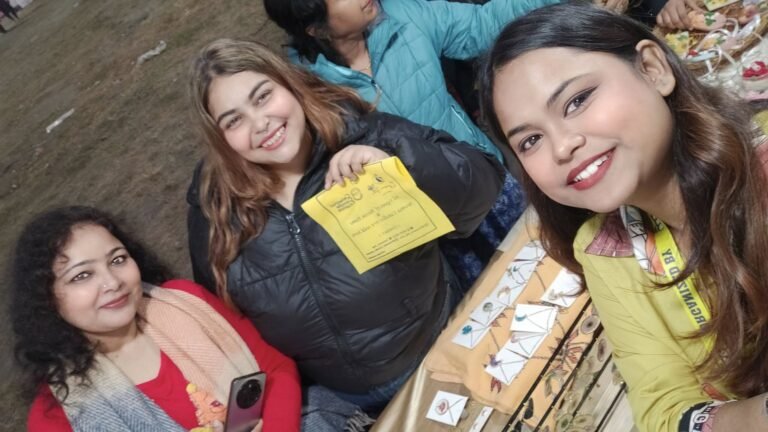 Three smiling women at an outdoor market stall, with handmade resin jewelry and keepsakes displayed on a table.