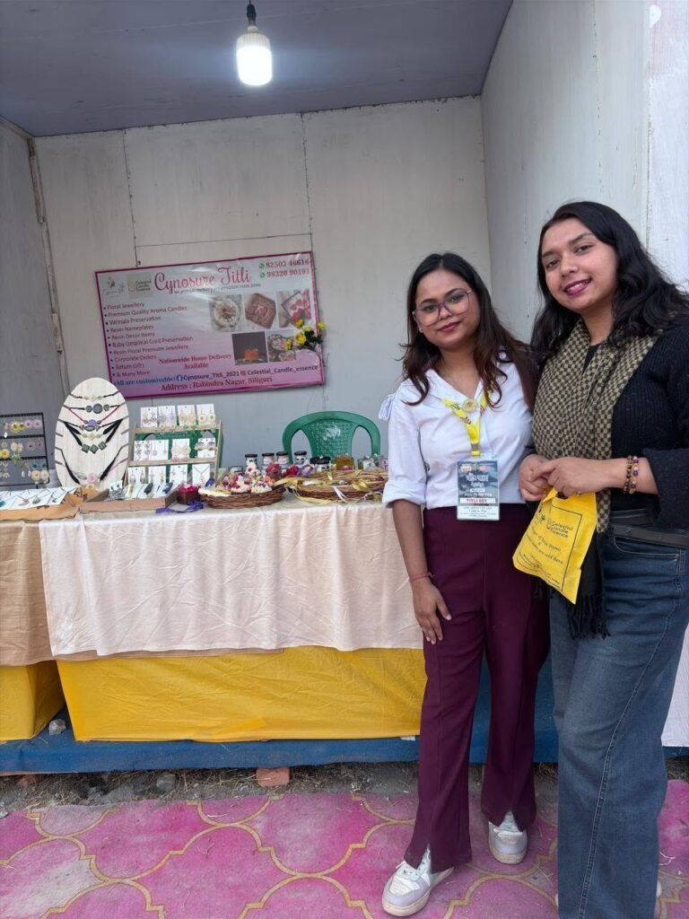 Two women smiling at their Cynosure Titli stall, displaying handmade resin jewelry, decor, and candles at a market.