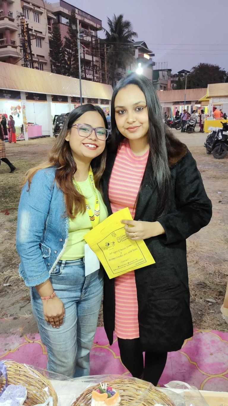Two smiling women at an outdoor market stall, one holding a yellow Cynosure Titli bag, with decorative items in baskets below