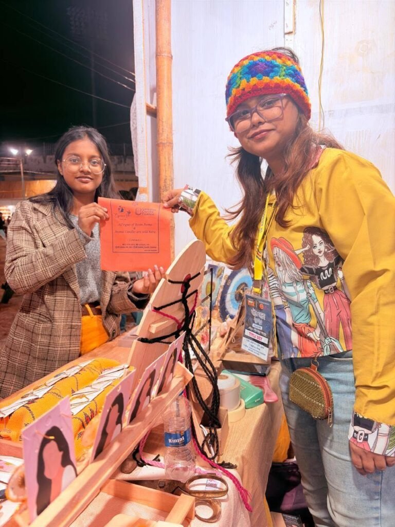 Two women at a market stall holding an orange sign that reads 'All types of Resin Items & Aroma Candles are sold here'.