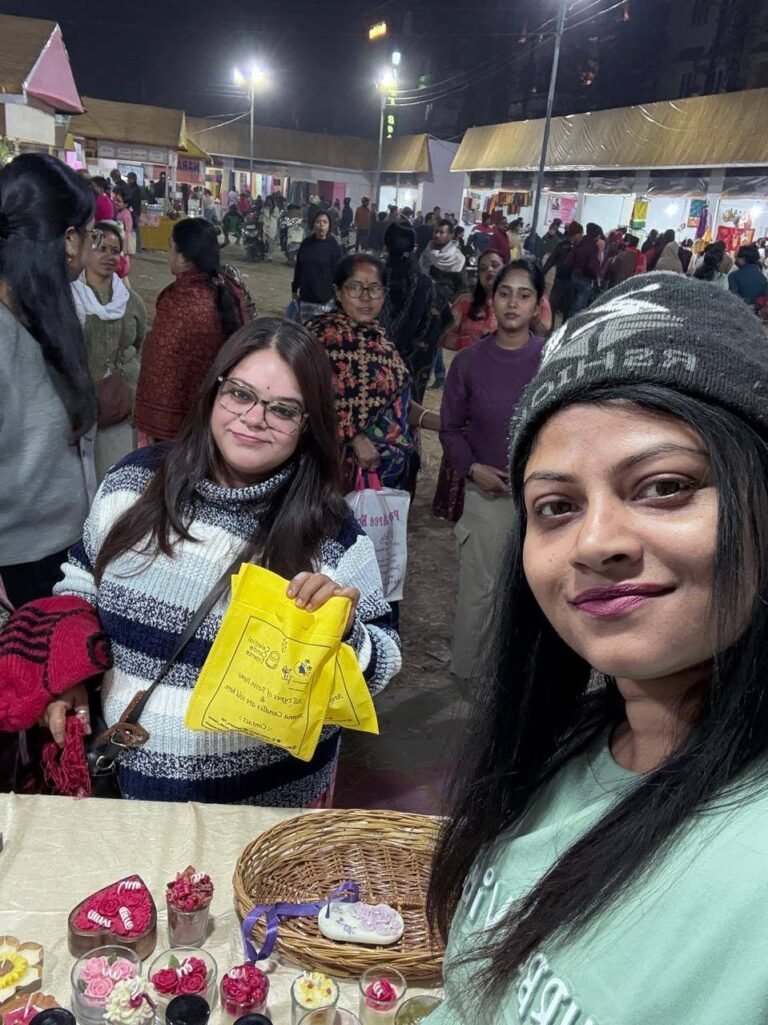 Smiling women at a night market stall with handmade scented candles and resin keepsakes, a yellow bag, and a bustling crowd.