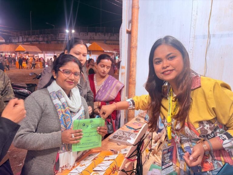 Two women smiling at a market stall with handmade products, one holding a green flyer, at an evening event.