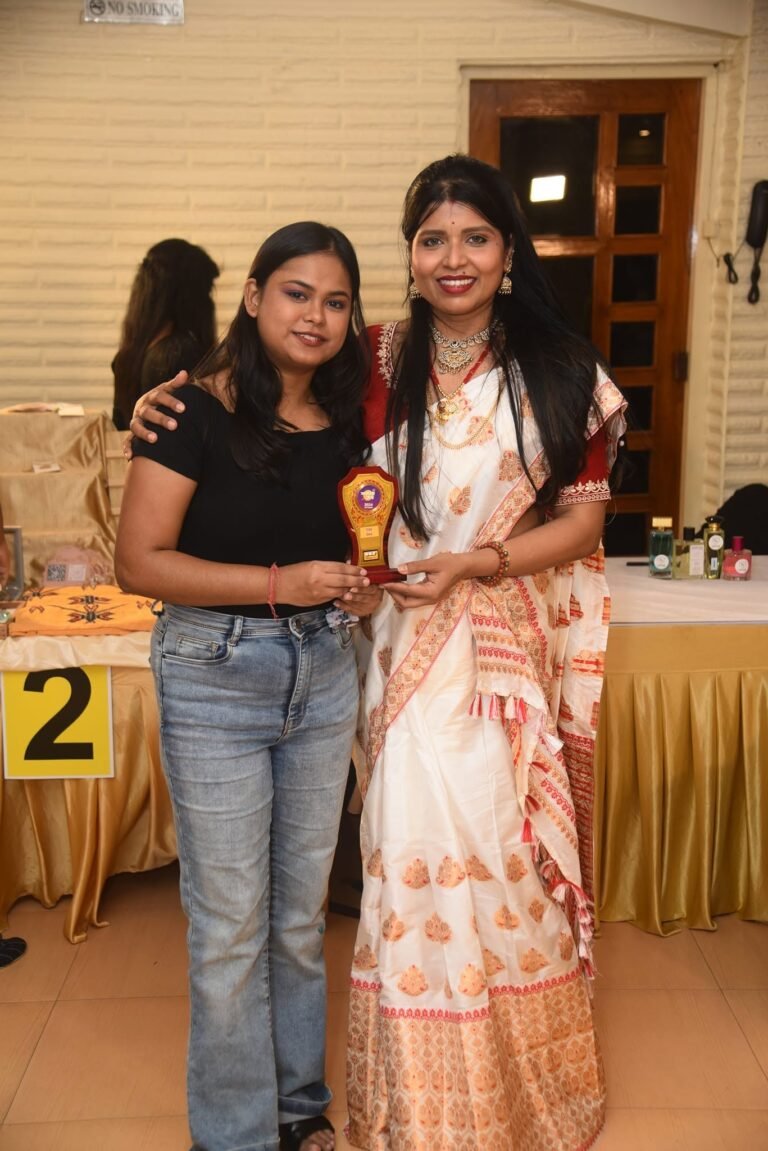 Two women smiling, one in a white and red saree, the other in jeans holding a wooden trophy, at an indoor event.