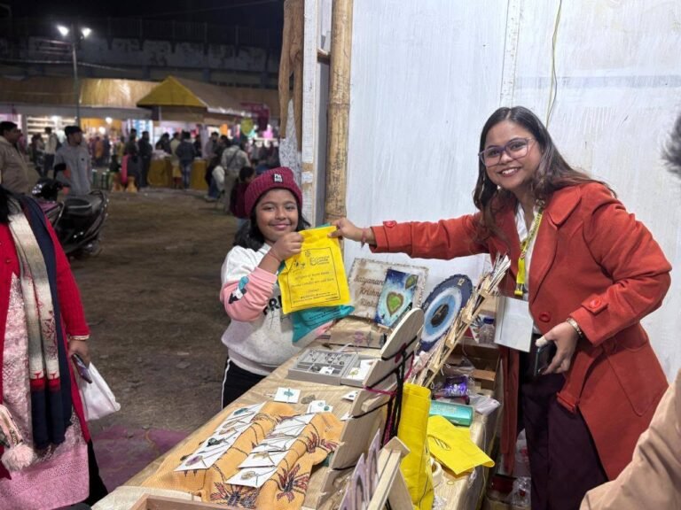 A smiling woman in an orange coat hands a yellow bag to a young girl at an outdoor market stall featuring handmade resin jewe