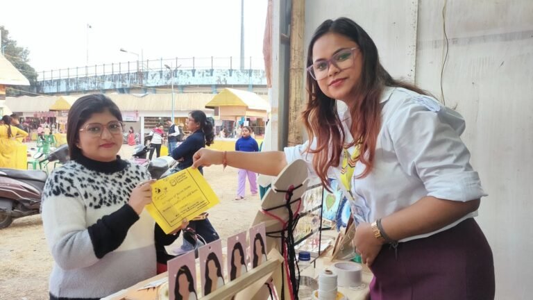 Two women at an outdoor market stall. One holds a yellow flyer, the other reaches out, with handmade earrings on display.
