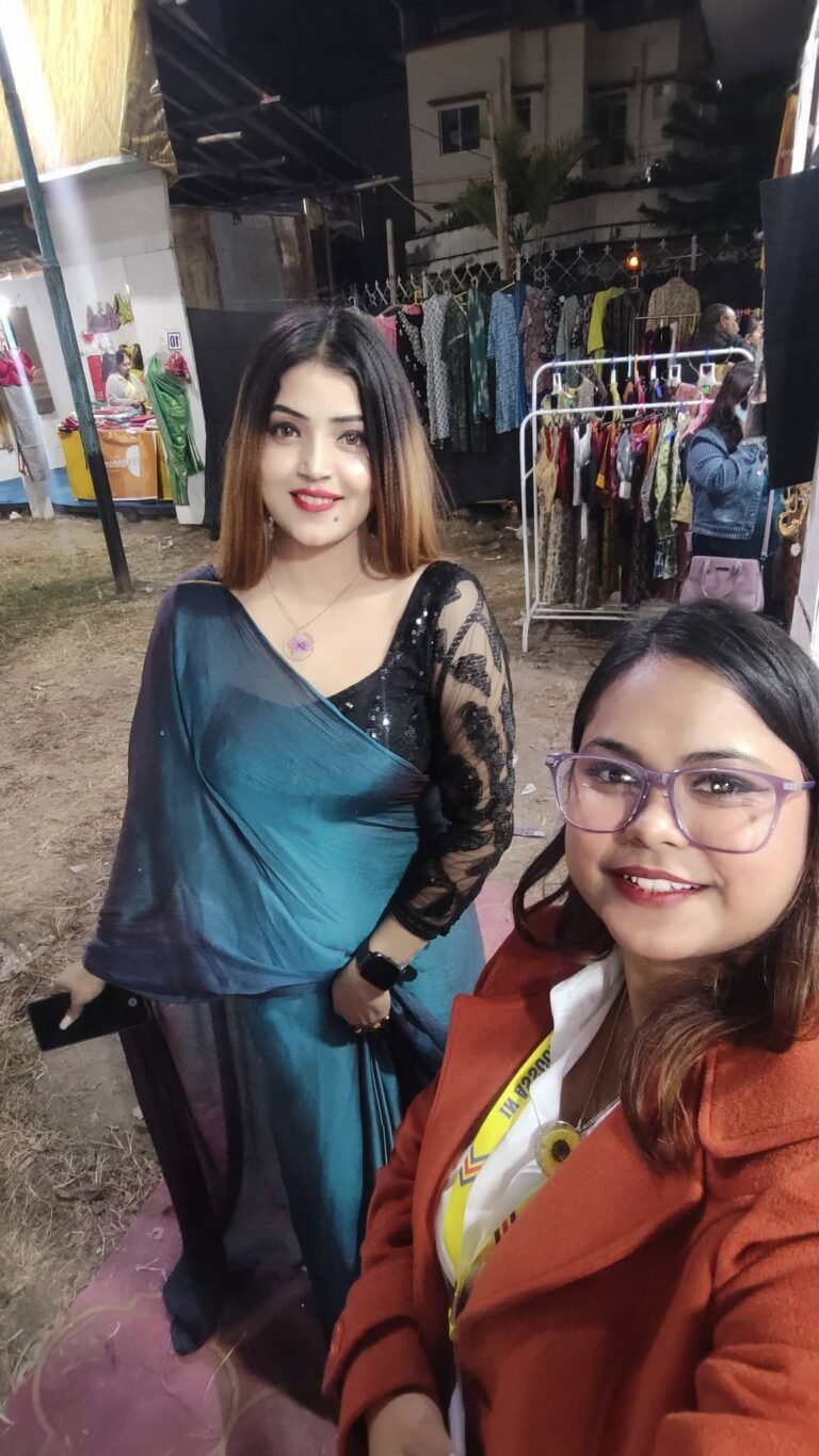 Two women smiling at a market stall. One wears a teal saree and floral pendant, the other an orange coat and glasses.