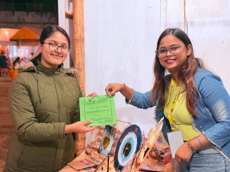 Two smiling women, one in a green jacket and one in a denim jacket, holding a green flyer at a stall with handmade items.