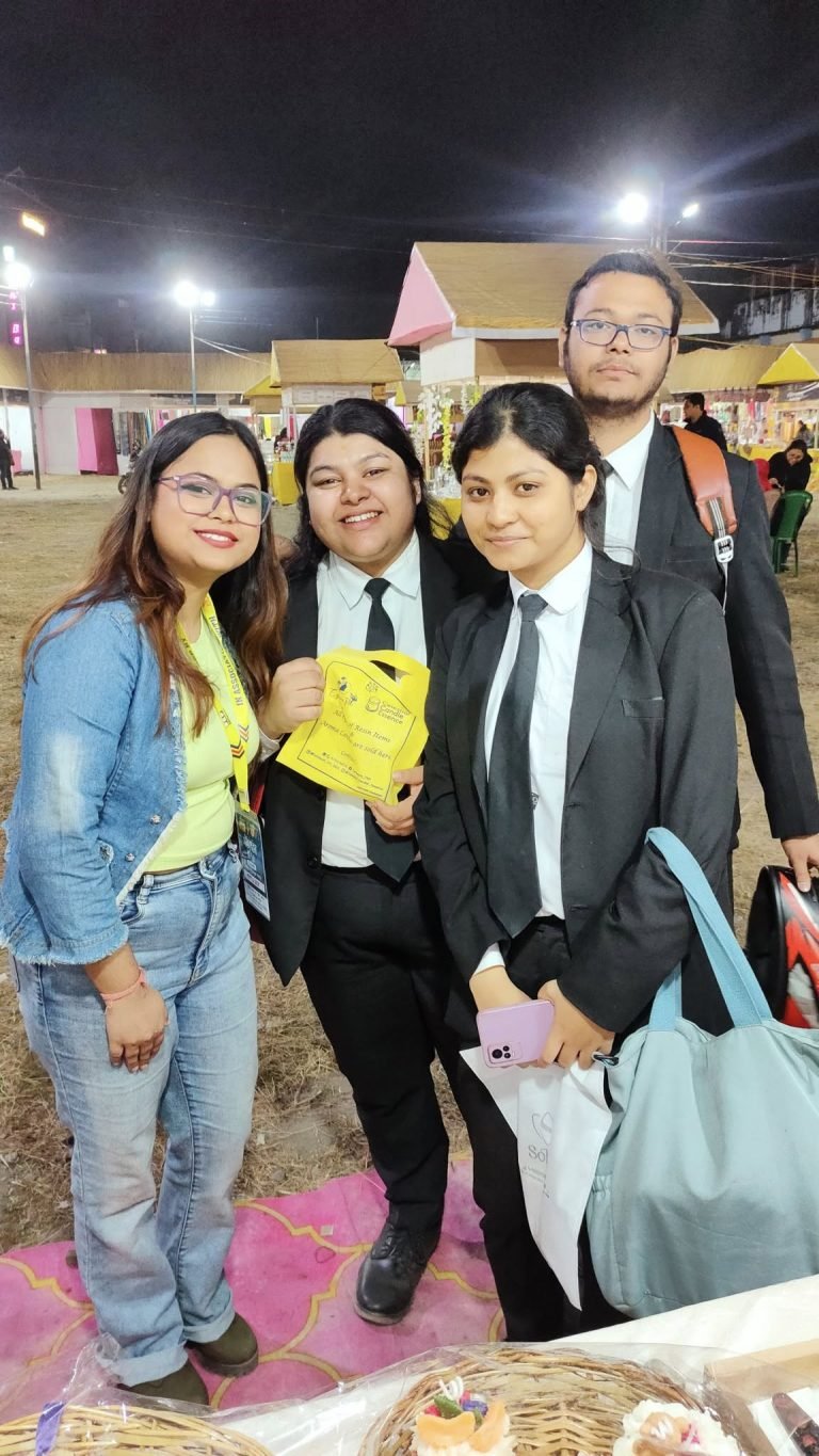 Four people, three in black suits and one in a denim jacket, smiling at an outdoor night market with stalls and lights.