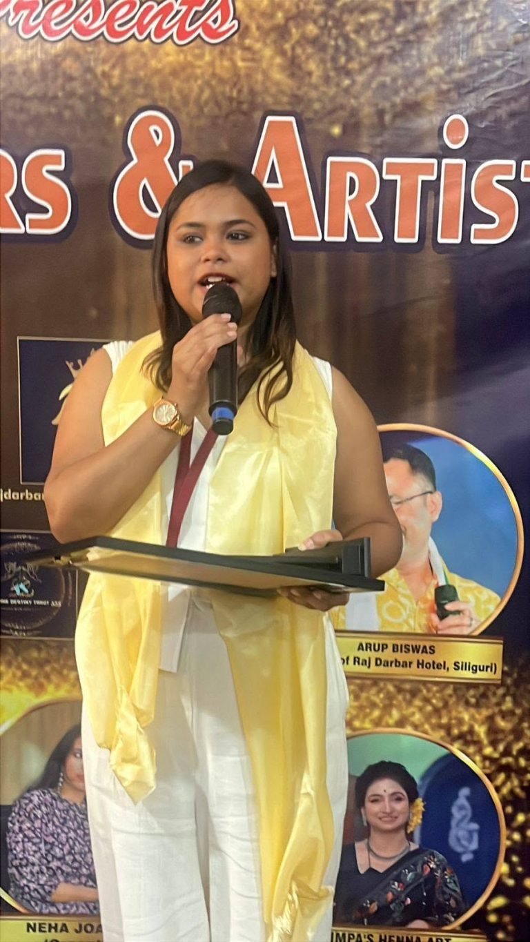 A woman in a white outfit and yellow stole speaks into a microphone, holding a black award plaque on stage.