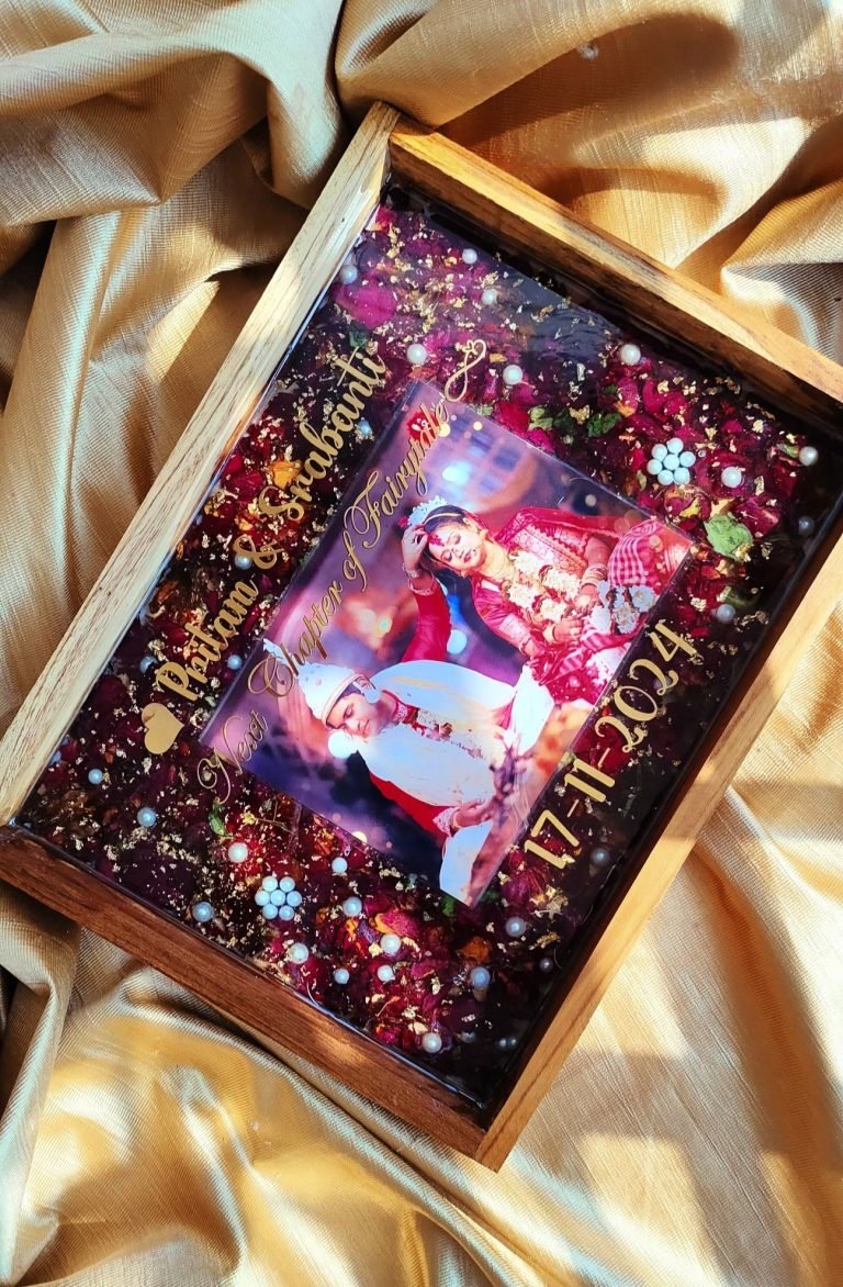 A custom resin keepsake tray with a wedding photo, red rose petals, gold flakes, pearls, and gold text on a golden fabric.