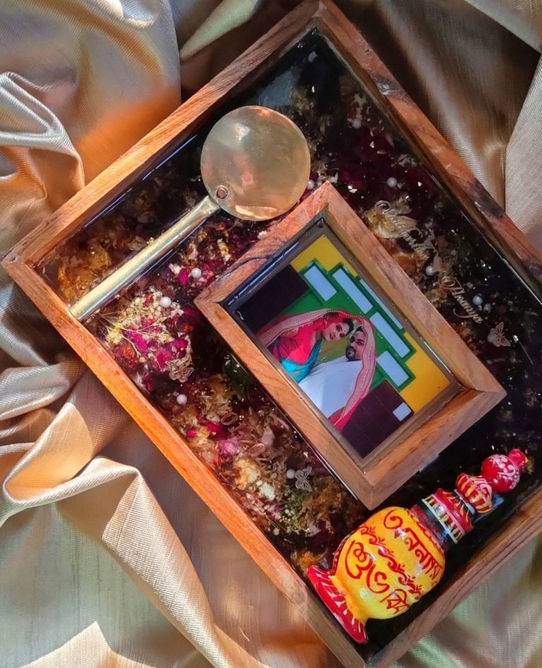 Warm-toned resin keepsake tray with dried flowers, a framed wedding photo, a golden spoon, and a decorative Bengali pot.