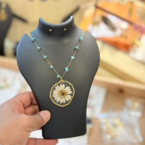 Gold-toned necklace with a white dried daisy resin pendant and turquoise beads, held on a black display stand.