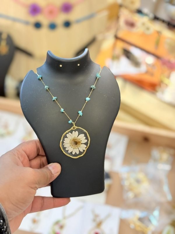 Gold-toned necklace with a white dried daisy resin pendant and turquoise beads, held on a black display stand.