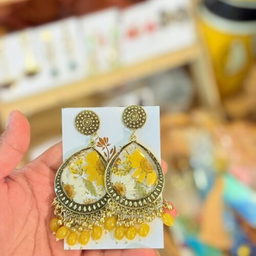 Hand holding golden teardrop earrings with preserved yellow and white flowers and yellow beads.
