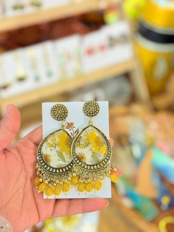 Hand holding golden teardrop earrings with preserved yellow and white flowers and yellow beads.