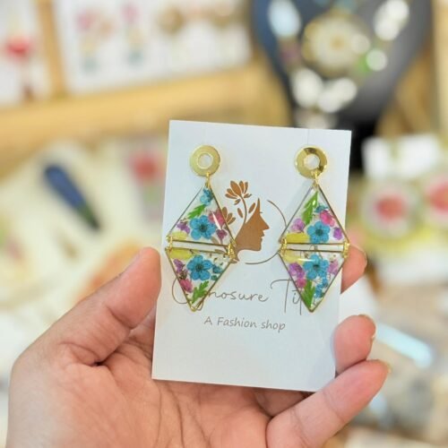 Hand holding diamond-shaped resin earrings with gold frames and preserved blue and purple flowers on a white display card.