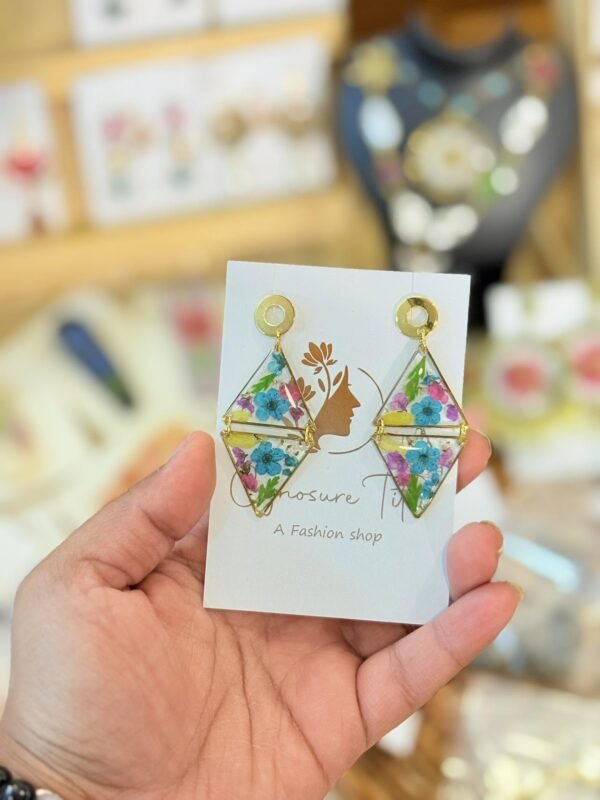 Hand holding diamond-shaped resin earrings with gold frames and preserved blue and purple flowers on a white display card.