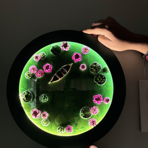 Round glowing green resin art featuring a boat, pink flowers, and lily pads, held by two hands.