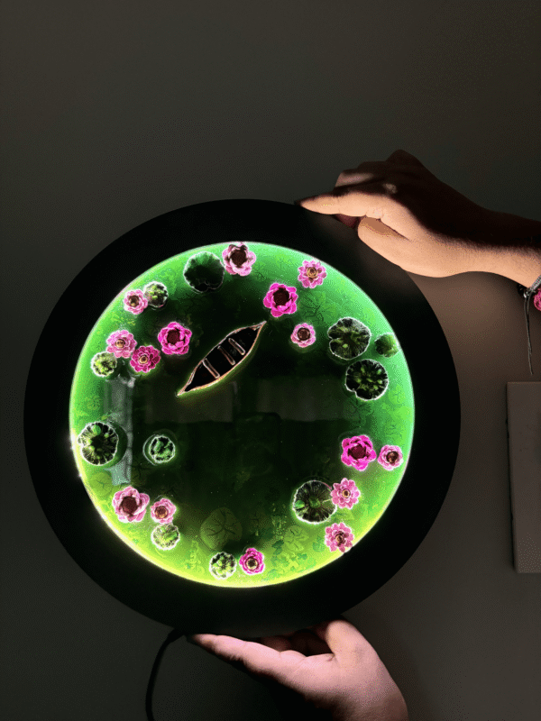 Round glowing green resin art featuring a boat, pink flowers, and lily pads, held by two hands.