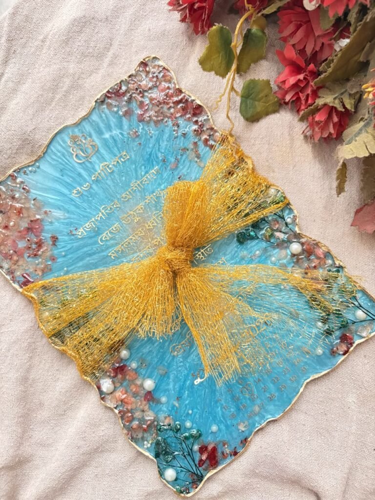Light blue resin keepsake with gold Bengali script, dates, crushed stones, pearls, and a gold mesh bow on beige fabric.