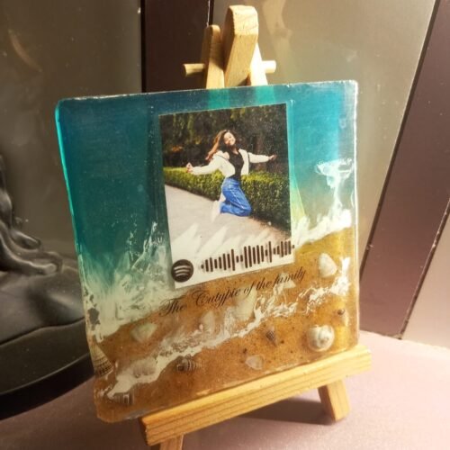 Resin keepsake with beach design, featuring a photo of a jumping woman, Spotify code, and text, displayed on a wooden easel.
