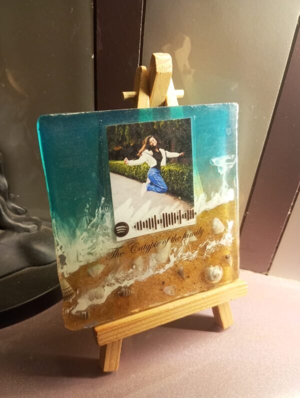 Resin keepsake with beach design, featuring a photo of a jumping woman, Spotify code, and text, displayed on a wooden easel.