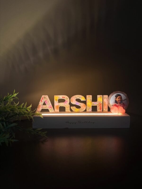 Illuminated resin nameplate "ARSHI" with colorful dried flowers, a child's photo, and glowing pink hearts on a "Happy Birthda