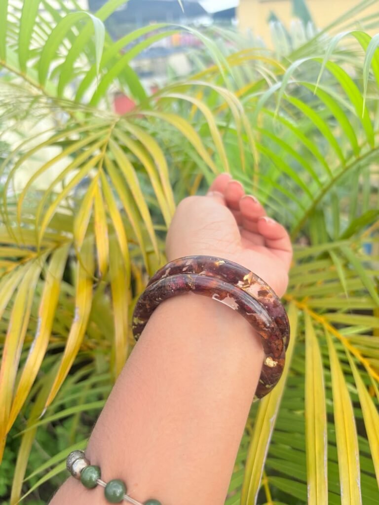 Arm wearing two dark red resin bangles with gold flakes, against vibrant green and yellow palm leaves.