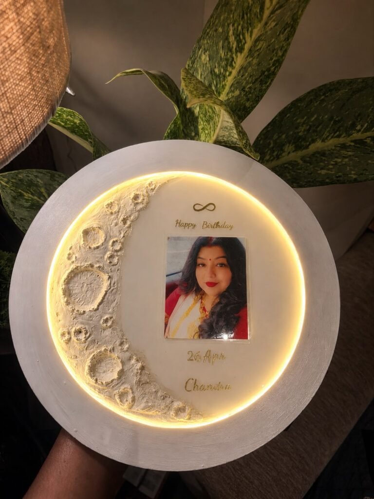 Round white illuminated moon-themed frame with a photo of a woman, 'Happy Birthday' and '26 Apr Chandru' in gold text.