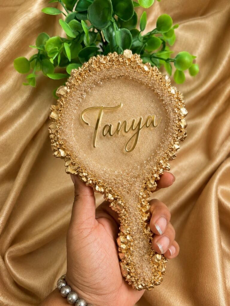 Hand holding a personalized golden resin mirror with "Tanya" in script, adorned with gold textured edges, against a golden fa